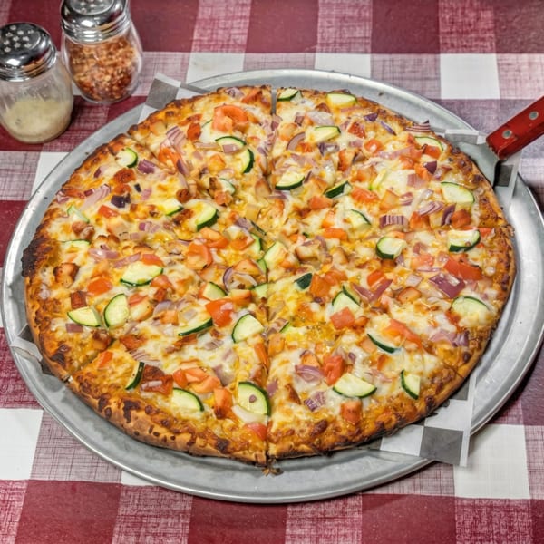 A whole pizza with zucchini, tomatoes, and red onions on a red and white checkered tablecloth.