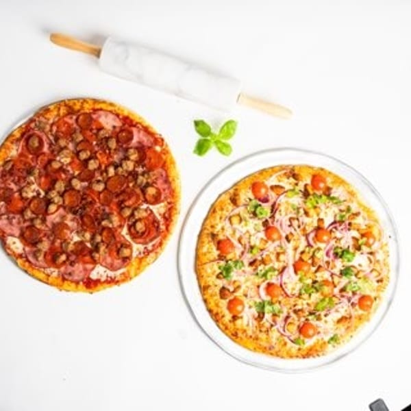 Two pizzas, one meat and one veggie, with a rolling pin and basil on a white surface.