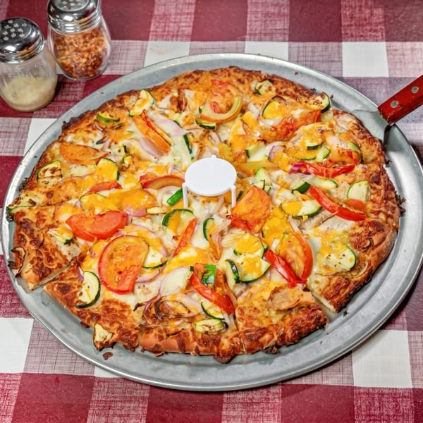 A large vegetable pizza on a metal pan with a red and white checkered tablecloth.
