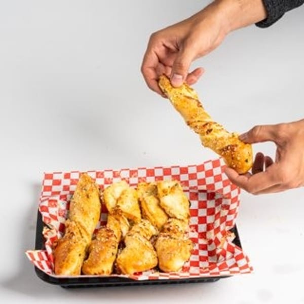 Hands holding a twisted breadstick above a tray of more breadsticks on a red and white checkered paper.
