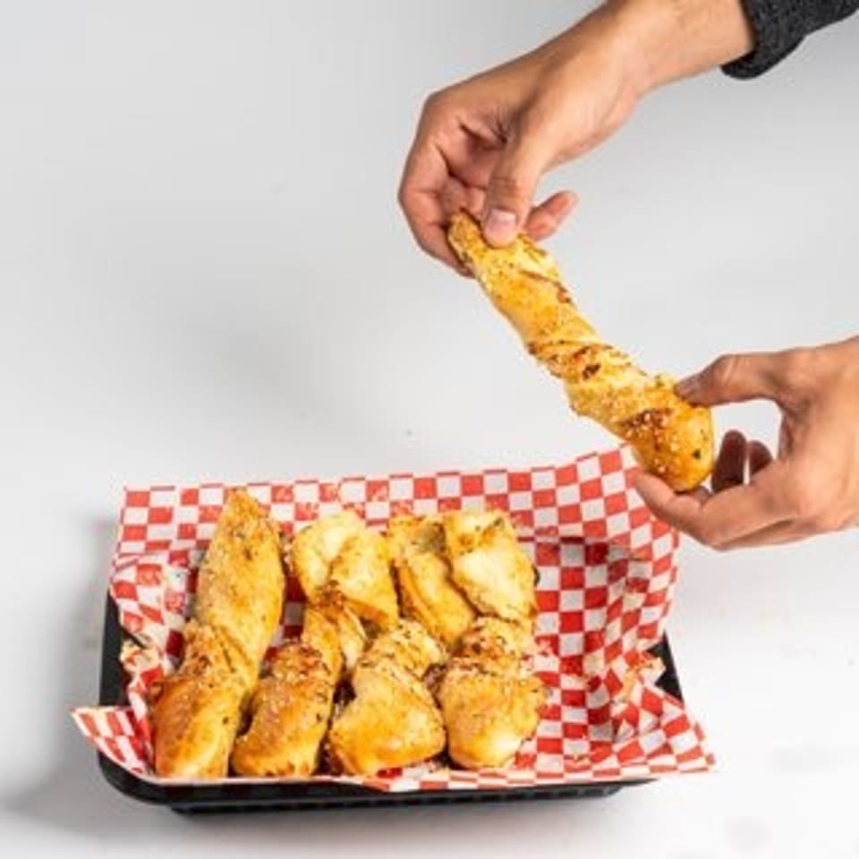 Hands holding a twisted breadstick above a tray of more breadsticks on a red and white checkered paper.