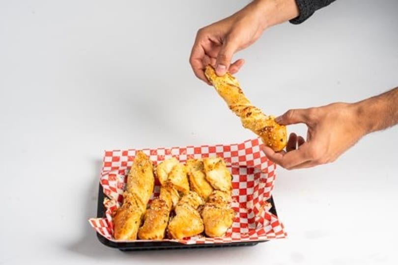 Hands holding a twisted breadstick above a tray of more breadsticks on a red and white checkered paper.