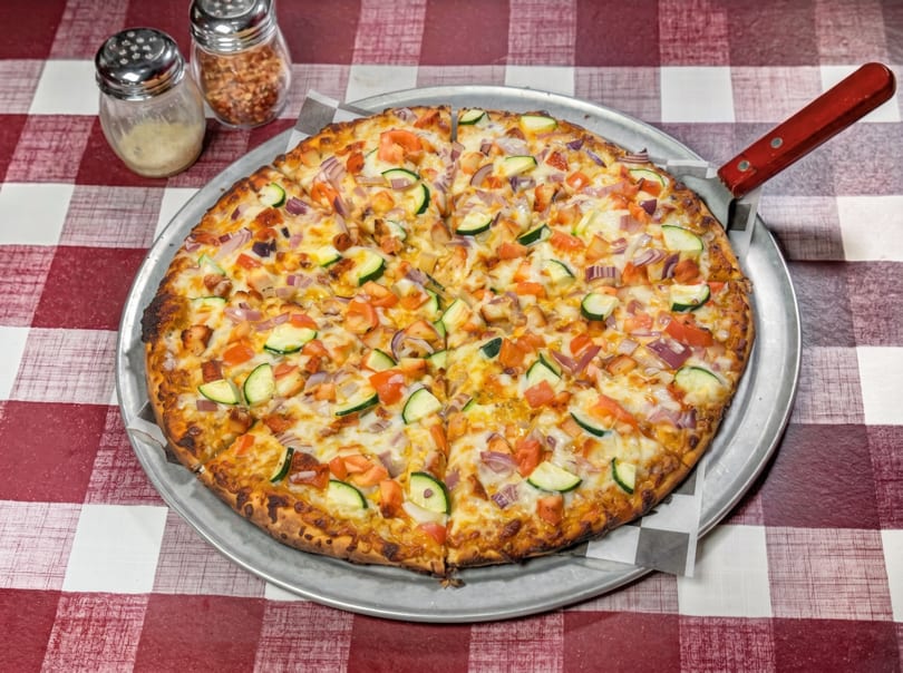 A freshly baked pizza with zucchini, tomatoes, and red onions on a red and white checkered tablecloth.