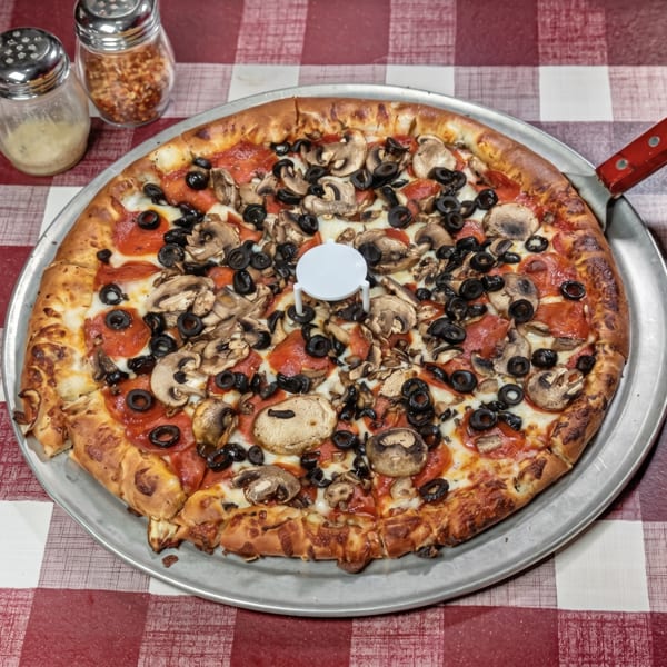A whole pepperoni, mushroom, and olive pizza on a metal pan with a red and white checkered tablecloth.