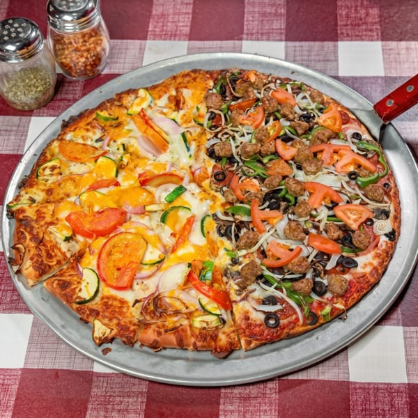 Half veggie, half meatball pizza on a metal pan with a red and white checkered tablecloth.