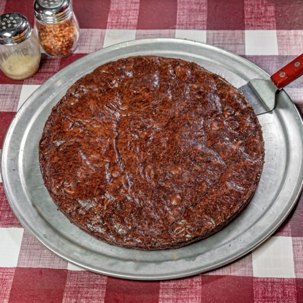 A large, round, dark brown brownie on a metal pizza pan with a red-handled spatula.