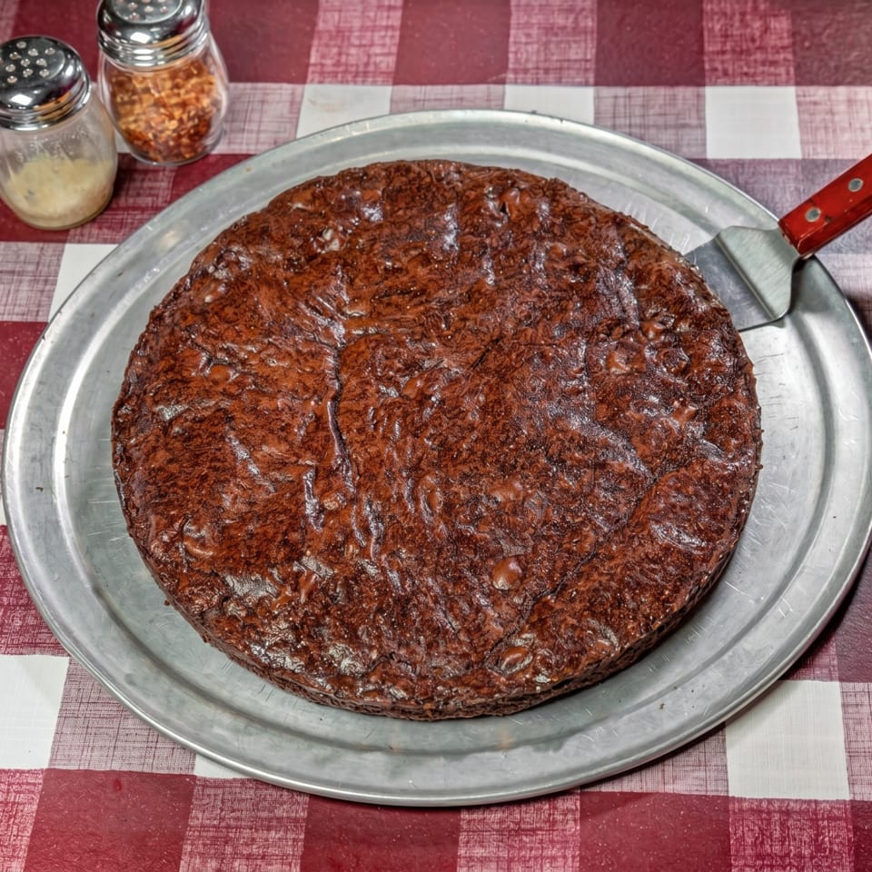 A large, round, dark brown brownie on a metal pizza pan with a red-handled spatula.