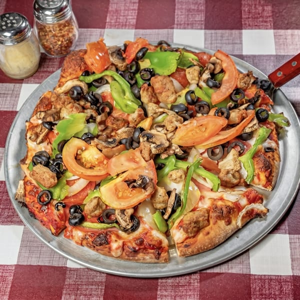 A whole pizza with sausage, mushrooms, olives, green peppers, and tomatoes on a checkered tablecloth.