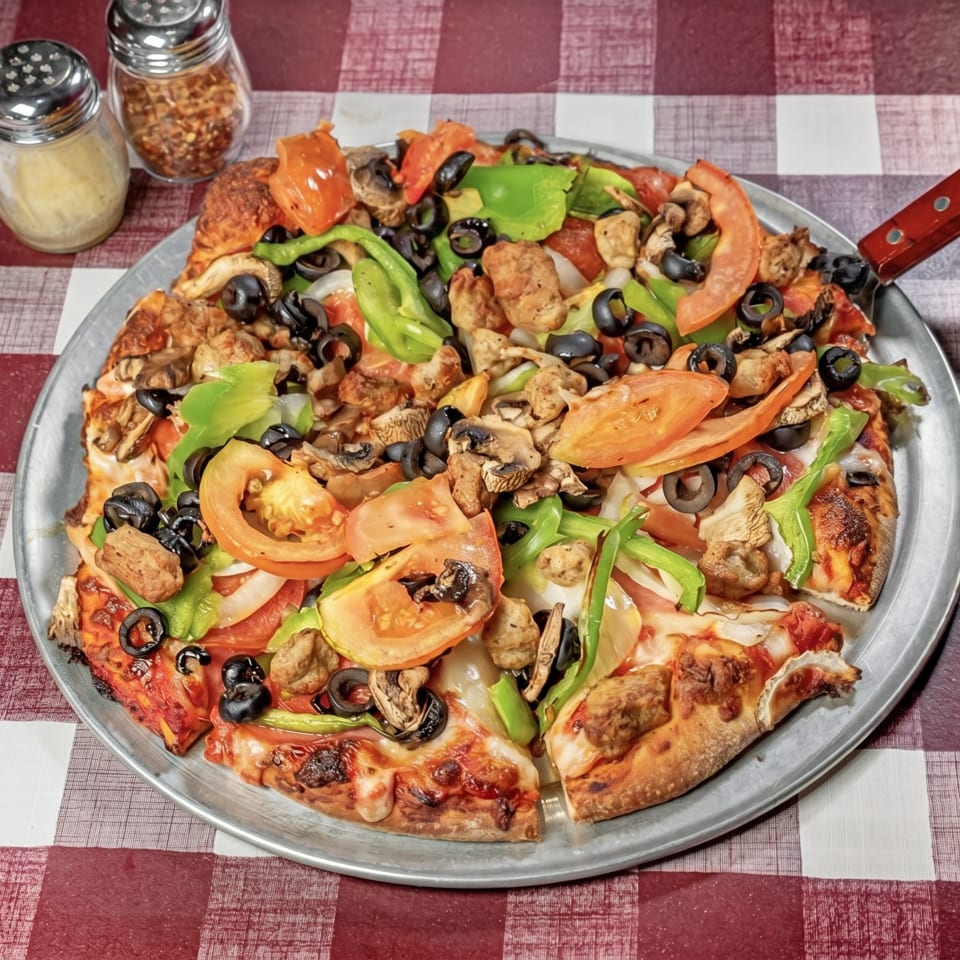 A whole pizza with sausage, mushrooms, olives, green peppers, and tomatoes on a checkered tablecloth.