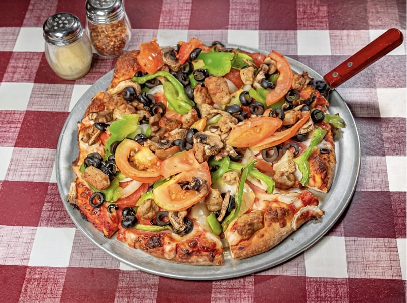 A whole pizza with sausage, mushrooms, olives, green peppers, and tomatoes on a checkered tablecloth.