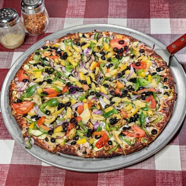 A large, colorful vegetable pizza on a metal pan with a server, on a red and white checkered tablecloth.