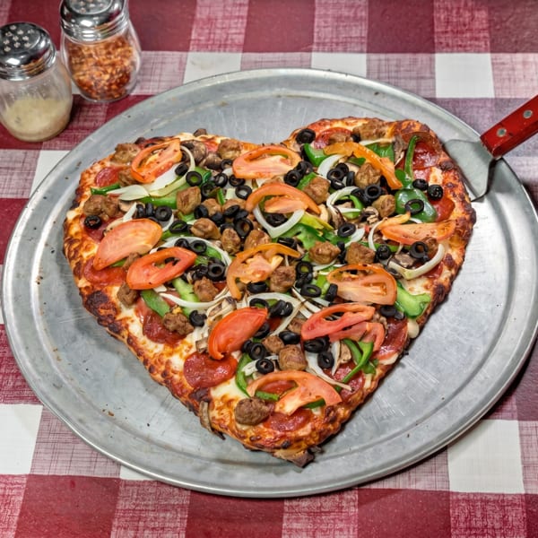 Heart-shaped pizza with pepperoni, sausage, olives, peppers, onions, and tomatoes on a metal tray.