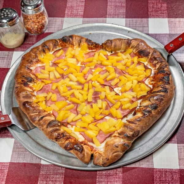 Heart-shaped pizza with ham and pineapple on a metal tray, with shakers on a red and white checkered tablecloth.