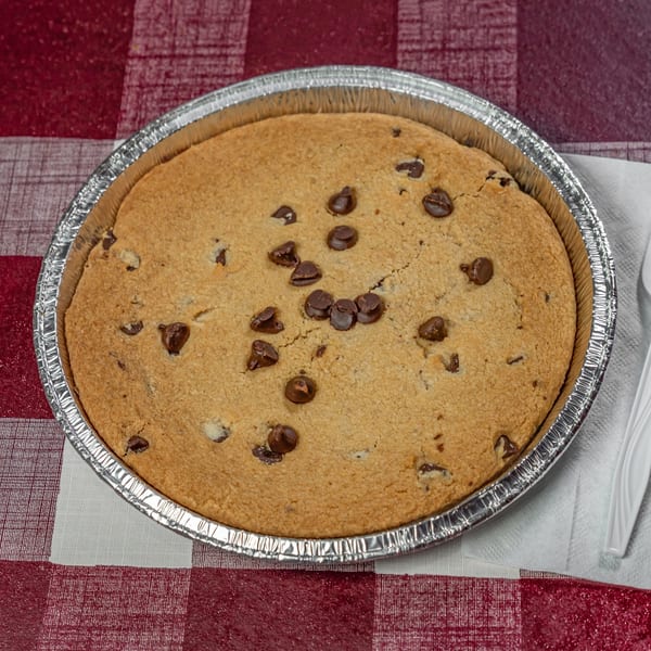 A large chocolate chip cookie in a foil pan with a white plastic fork on a red and white checkered tablecloth.